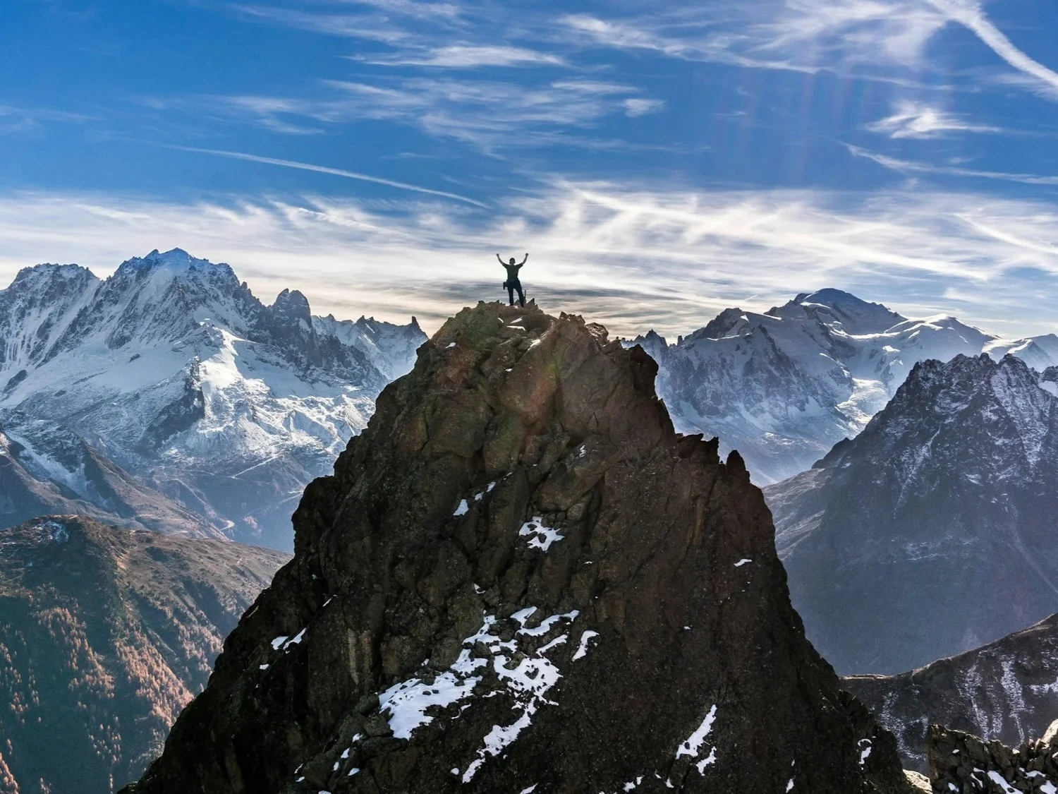 A person standing triumphantly at the summit of a mountain ridge with snow-capped peaks behind
