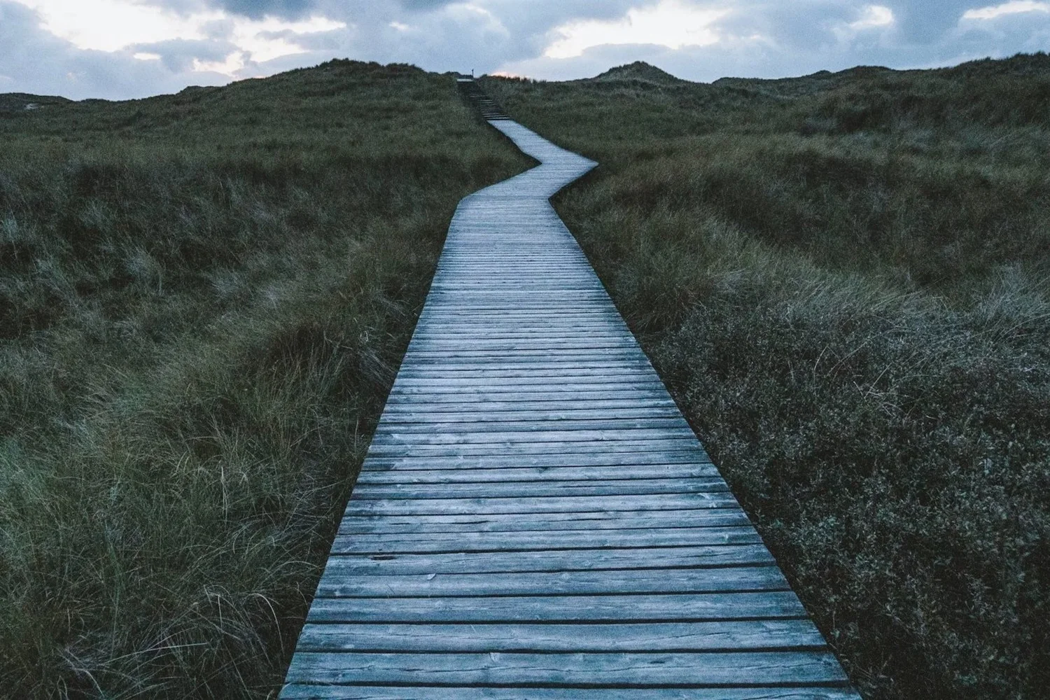 A winding wooden pathway through grassy dunes under a cloudy sky